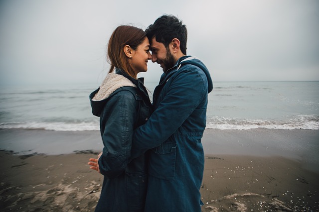 Žena a muž v dlhým modrých bundách stoja na pláži oproti sebe, dotýkajú sa tvárami.jpg